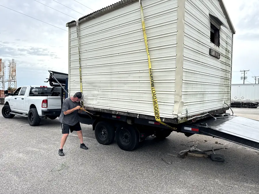A shed strapped down on the trailer with a crew member working the straps